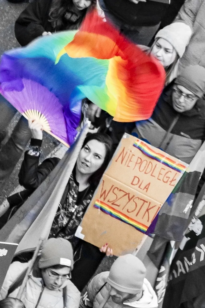Protest crowd beneath rainbow flags and a cardboard sign